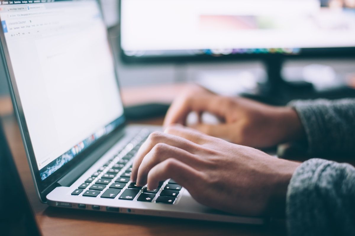 Hands typing on a laptop keyboard with a document open, a large monitor blurred behind