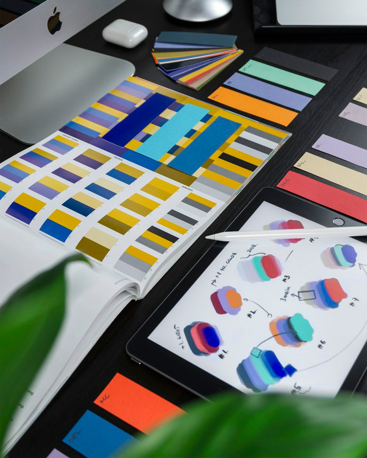 Desk covered with paper color swatches, an open color palette book, and a tablet with color sketches