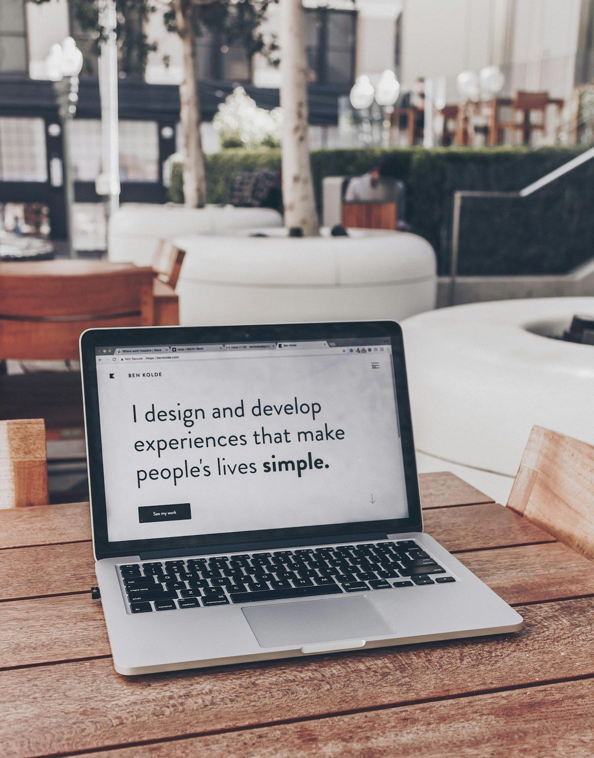 Laptop on an outdoor wooden cafe table showing a minimalist portfolio site with a short headline
